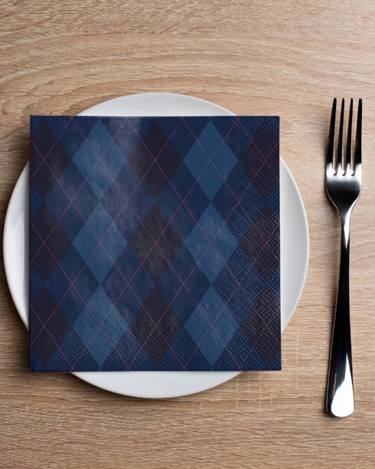 Blue Faire Argyle cocktail napkin by Bungalow on white plate with silverware, set on a wooden dining table.