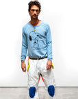 A man stands against a plain white background, wearing the Free City BasicGoodness Schulz Raglan by sparrow, LLC— a light blue unisex sweatshirt featuring Snoopy hugging Woodstock—paired with white pants with blue knee patches and yellow accents.