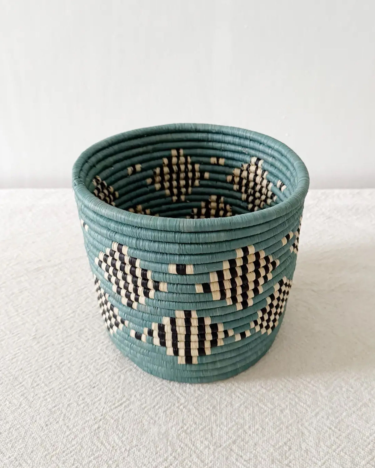 The Blue Storage Plant Basket: Mifuti by Faire features teal, black, and natural geometric patterns. Handwoven in Rwanda, this unique basket planter adds an authentic touch of African decor to any room.