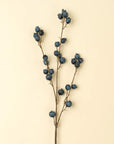 The Indaba Blueberry Branch features multiple stems with clusters of dried blue berries, set against a beige background—ideal as accent decor or combined with faux flowers.