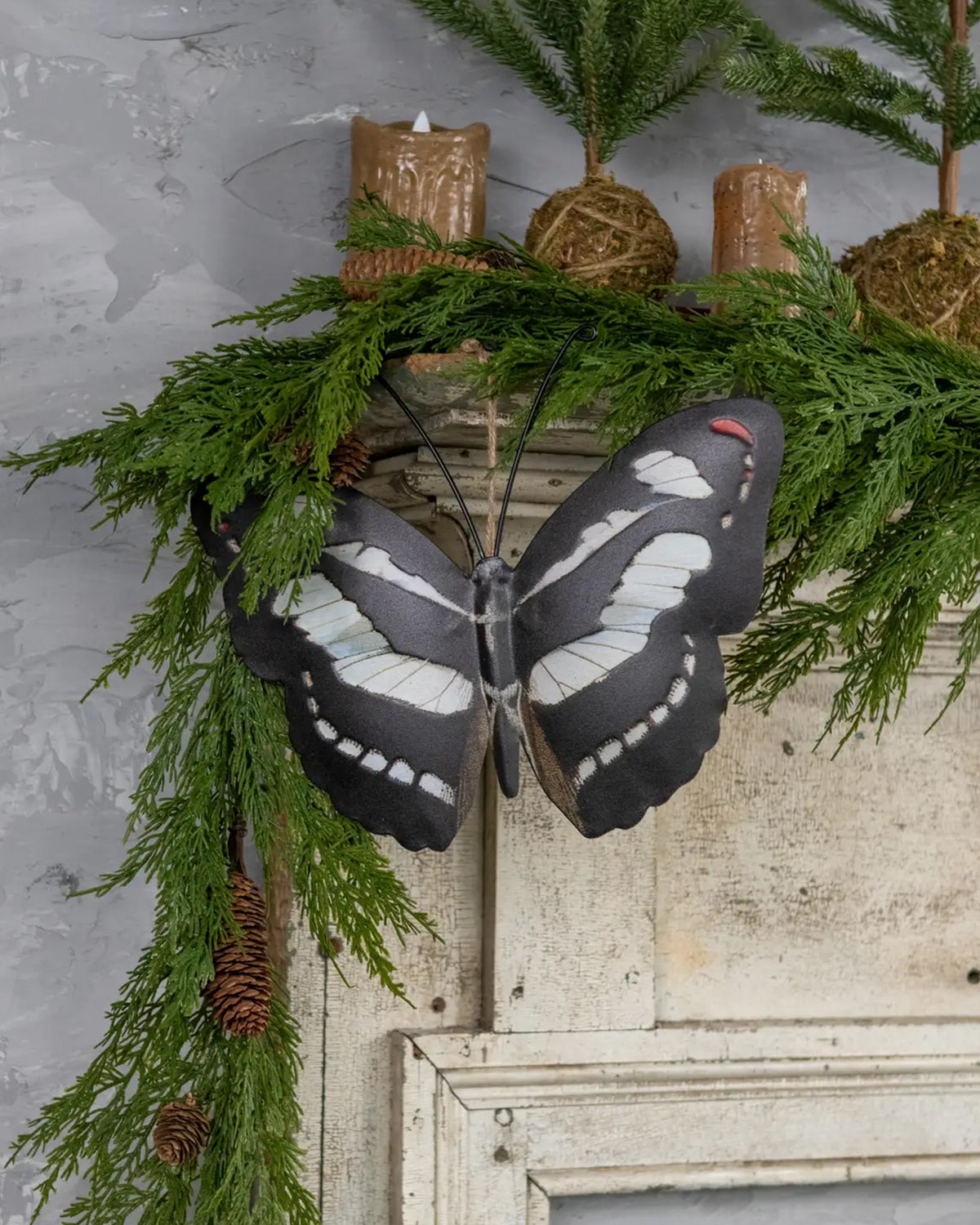 Black and white butterfly décor by Faire tops pine garland on a white mantel, styled with brown candles.