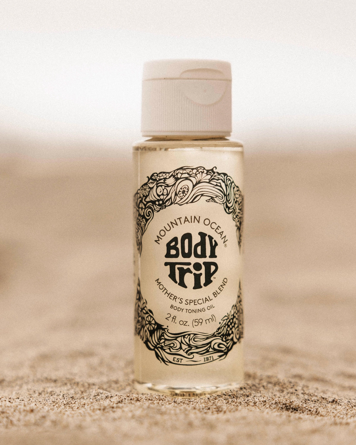 A small bottle of Faire Body Trip Travel Size skin toning oil stands upright on sand, displaying ornate black designs and text on a clear label. The softly blurred background highlights this natural skincare essential.