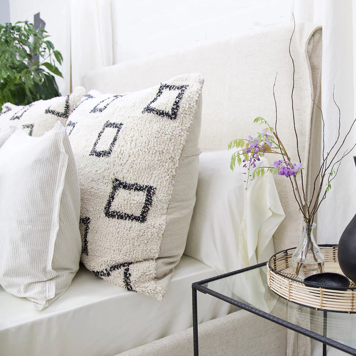 A cozy bed with a POM POM Bowie Big Pillow and neutral bedding sits beside a glass nightstand holding a woven tray, vase with purple flowers, and black decor. A green leafy plant is visible in the background.