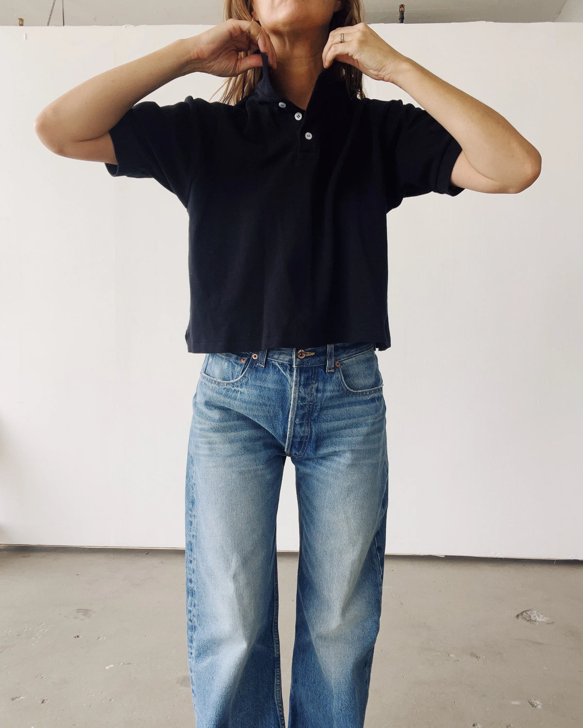 A person stands indoors against a white wall, wearing Hey Gang’s The Boxy Polo in black cotton pique with blue jeans. Their hands are raised to their neck, and their face is partially out of the frame.