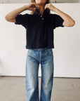 A person stands indoors against a white wall, wearing Hey Gang’s The Boxy Polo in black cotton pique with blue jeans. Their hands are raised to their neck, and their face is partially out of the frame.
