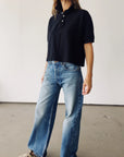 A person stands indoors against a plain white background, wearing Hey Gang's The Boxy Polo in black paired with high-waisted, wide-leg blue jeans. Their face is not visible.