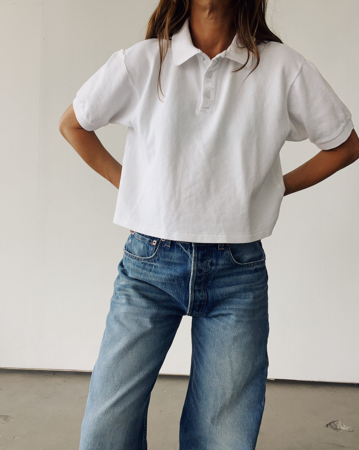 A person wearing Hey Gang’s The Boxy Polo and blue jeans stands hands-on-hips, facing forward against a plain light background, head partially out of frame.