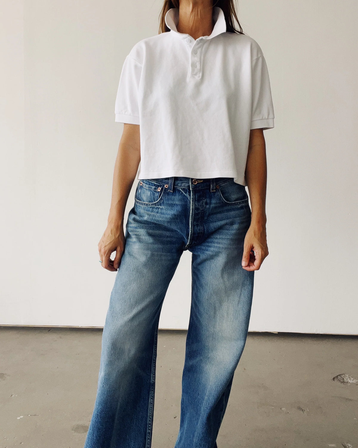 Against a plain background, a person wears Hey Gang's The Boxy Polo in white cotton pique with the collar popped and high-waisted, wide-leg blue jeans. Their face is not visible.