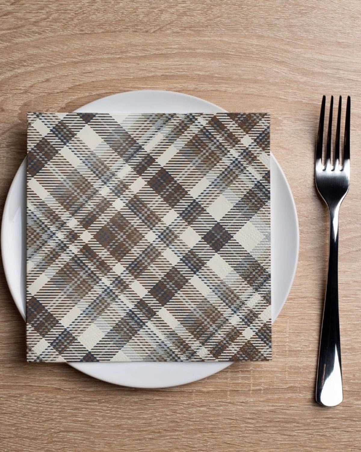Brown plaid cocktail napkin by Faire on white plate with fork, set on a wooden table.