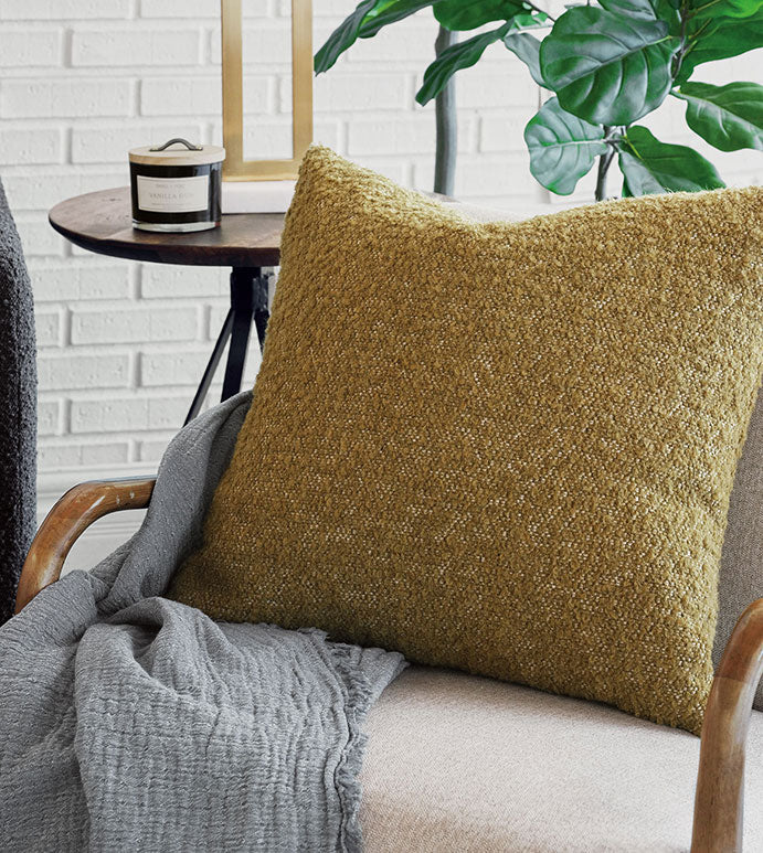 A cozy living room features the Eastern Accents Marl Mustard Pillow with a textured finish and invisible zipper, paired with a soft gray blanket, a round side table holding a candle, and a leafy green plant against a white brick wall.