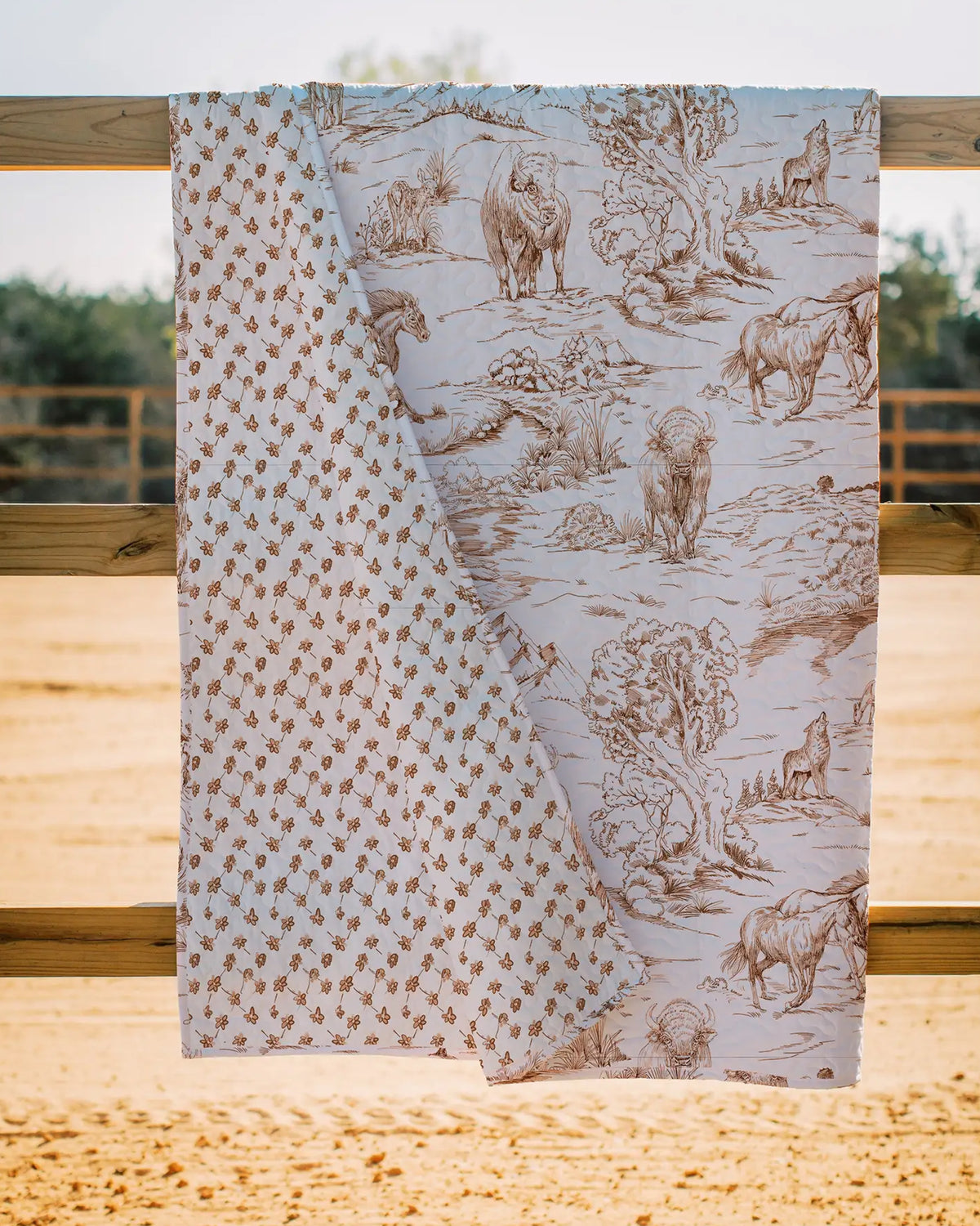 The Faire Buffalo Toile Brown Quilt is draped over a wooden fence, displaying brown wildlife and nature scenes on one side and a small repeating floral or animal print on the other. Set outdoors with sand and trees, it's ideal for cozy bedding.