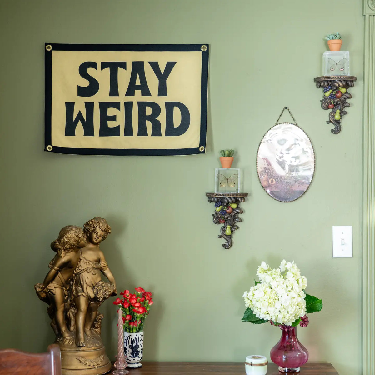 Stay Weird Camp Flag by Faire displayed on a green wall, adding playful style above floral and eclectic decor.