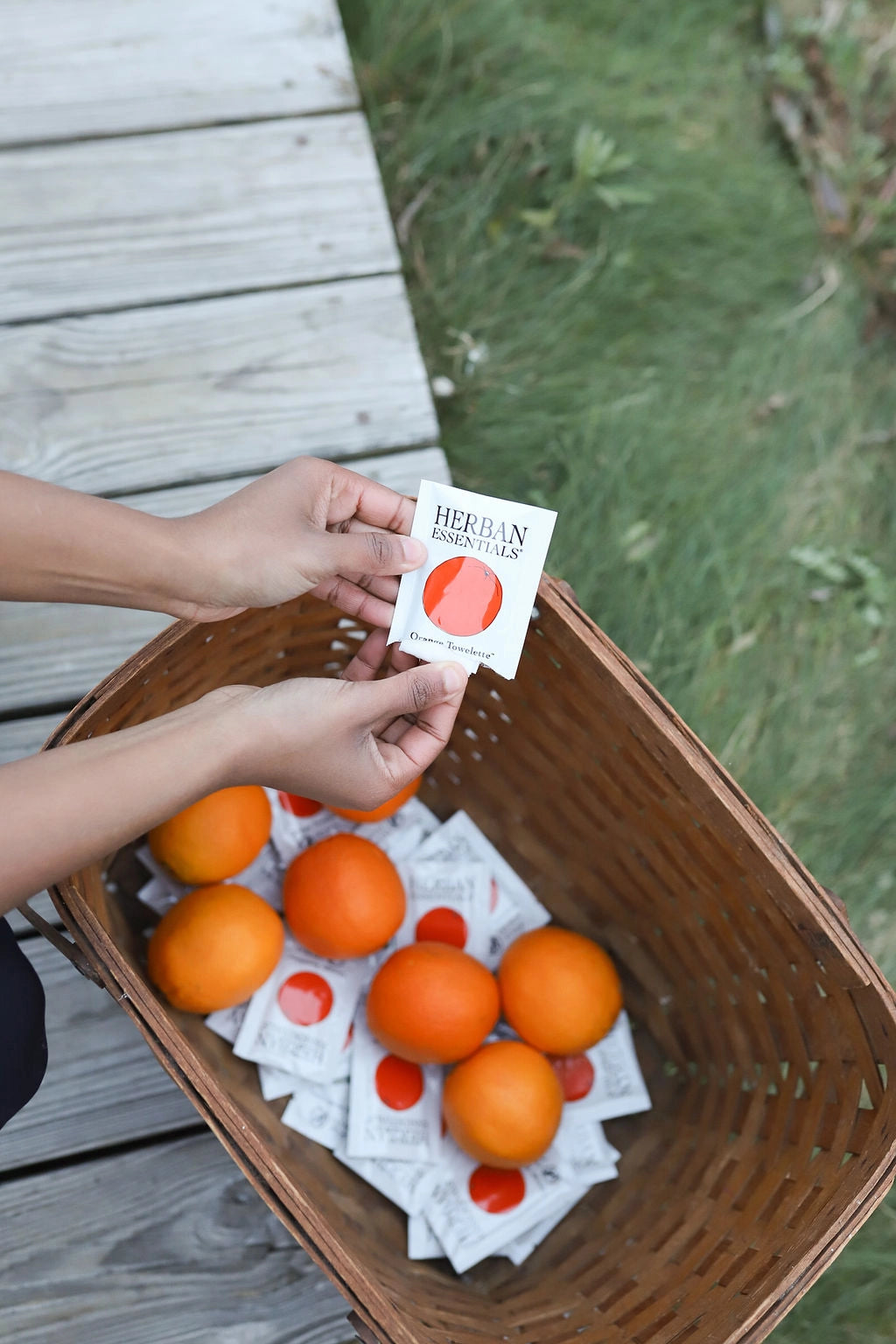 A person holds a Faire Orange Towelettes pack—20 individual essential oil wipes—above a basket of fresh oranges and wrapped wipes, all arranged on a wooden deck next to green grass.