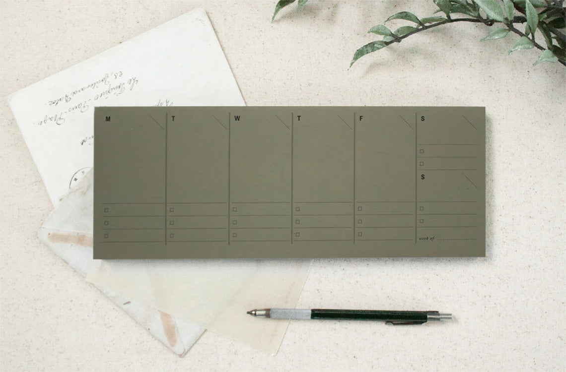 The Faire Weekly Planner Paper Notepad, a minimalist weekly organizer with columns for each day, is displayed on a cream surface next to vintage letters, a clear envelope, a black pen, and green leaves.