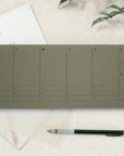 The Faire Weekly Planner Paper Notepad, a minimalist weekly organizer with columns for each day, is displayed on a cream surface next to vintage letters, a clear envelope, a black pen, and green leaves.