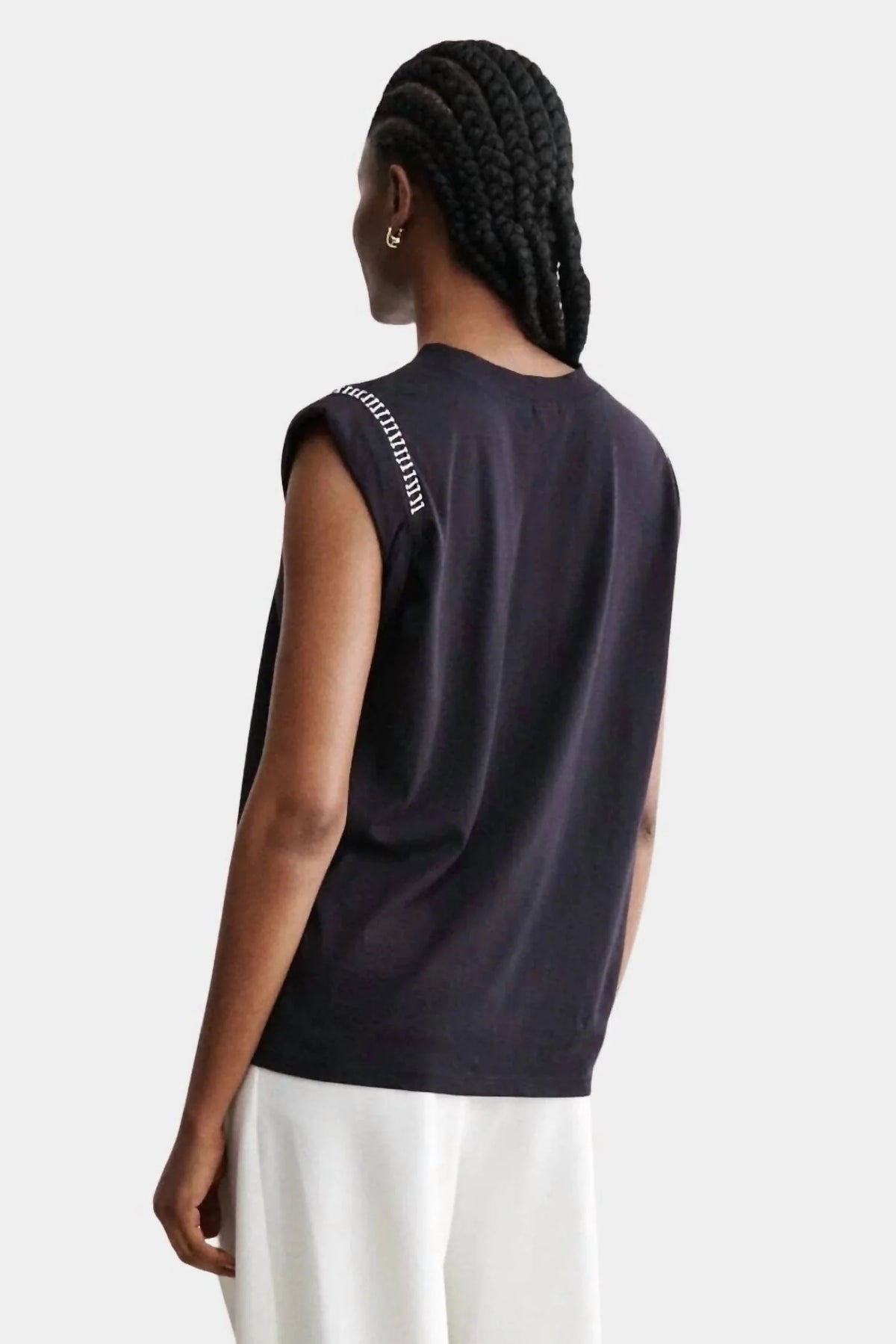 A person with braided hair is seen from behind in a sleeveless black Rachel Comey Miles Tee with shoulder text detail and white pants, standing against a plain light gray background for a minimalist look.