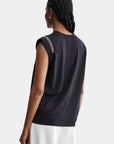 A person with braided hair is seen from behind in a sleeveless black Rachel Comey Miles Tee with shoulder text detail and white pants, standing against a plain light gray background for a minimalist look.
