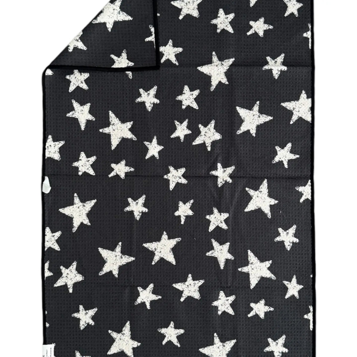 The Faire "Hand Towel : Into The Stars" is a black rectangular waffle weave microfiber towel with white stars of various sizes. A folded corner shows that the same star pattern appears on both sides.