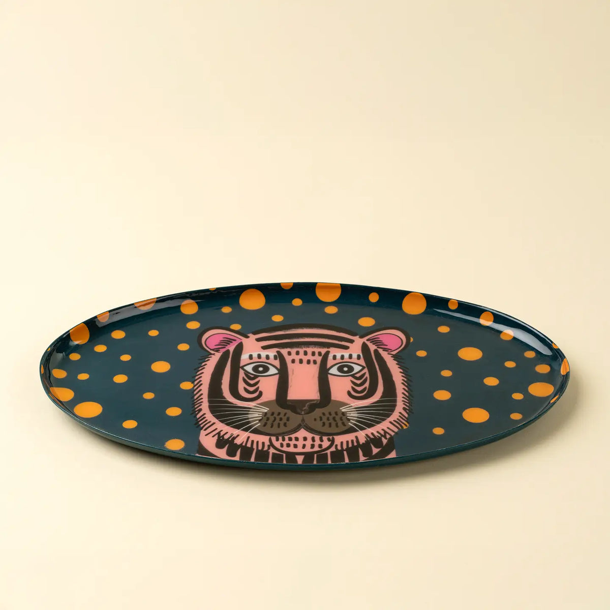 The Indaba Tiger Spot Tray features a dark background, orange polka dots, and a pink tiger face at the center, bringing a playful touch to your décor. Perfect as an accent piece when displayed on any plain surface.