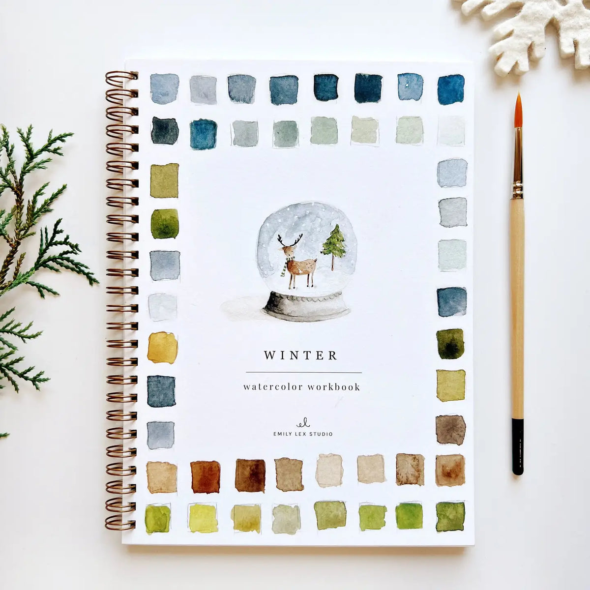 The spiral-bound Faire Watercolor Workbook, titled "Winter," showcases a snow globe with a deer, trees, and color swatches on the cover. Beside it are a pine sprig, paintbrush, and snowflake ornament set against a white background.