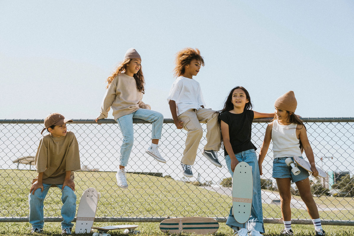 Five kids hang out by a chain-link fence on a sunny day. Two sit atop the fence while three stand or lean nearby, showing off their Faire Vintage Skateboards. All are dressed casually and enjoying time together.