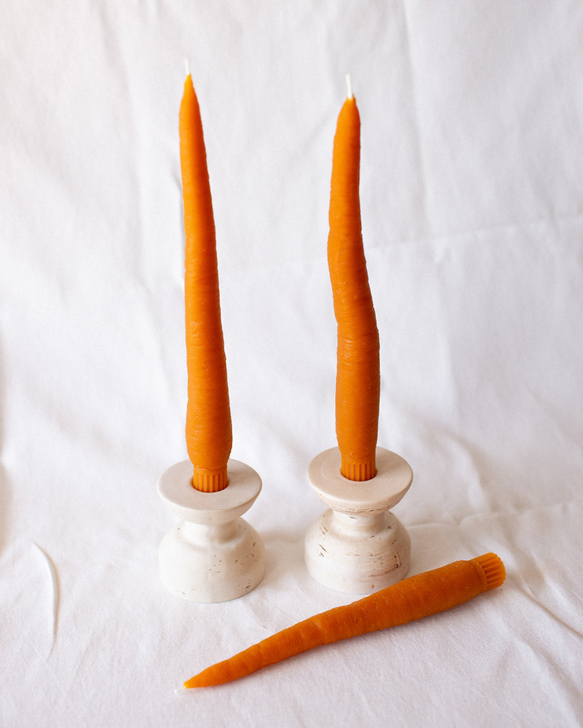 Three Faire Carrot Beeswax Candles are displayed: two upright in white holders and one lying down, all shown against a plain white background.
