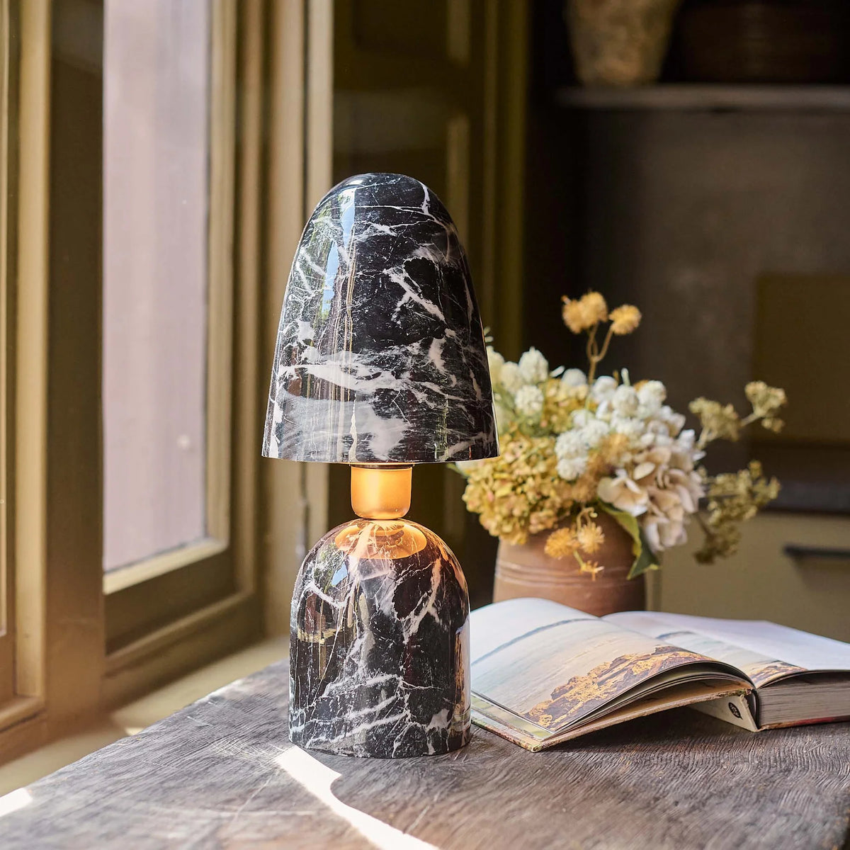 The Caswell Table Lamp by Abigail Ahern, with a black marble finish, illuminates a wooden desk set with an open book and dried flowers as sunlight pours in, adding modern accent lighting to the room.
