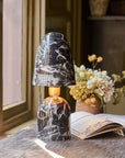 The Caswell Table Lamp by Abigail Ahern, with a black marble finish, illuminates a wooden desk set with an open book and dried flowers as sunlight pours in, adding modern accent lighting to the room.