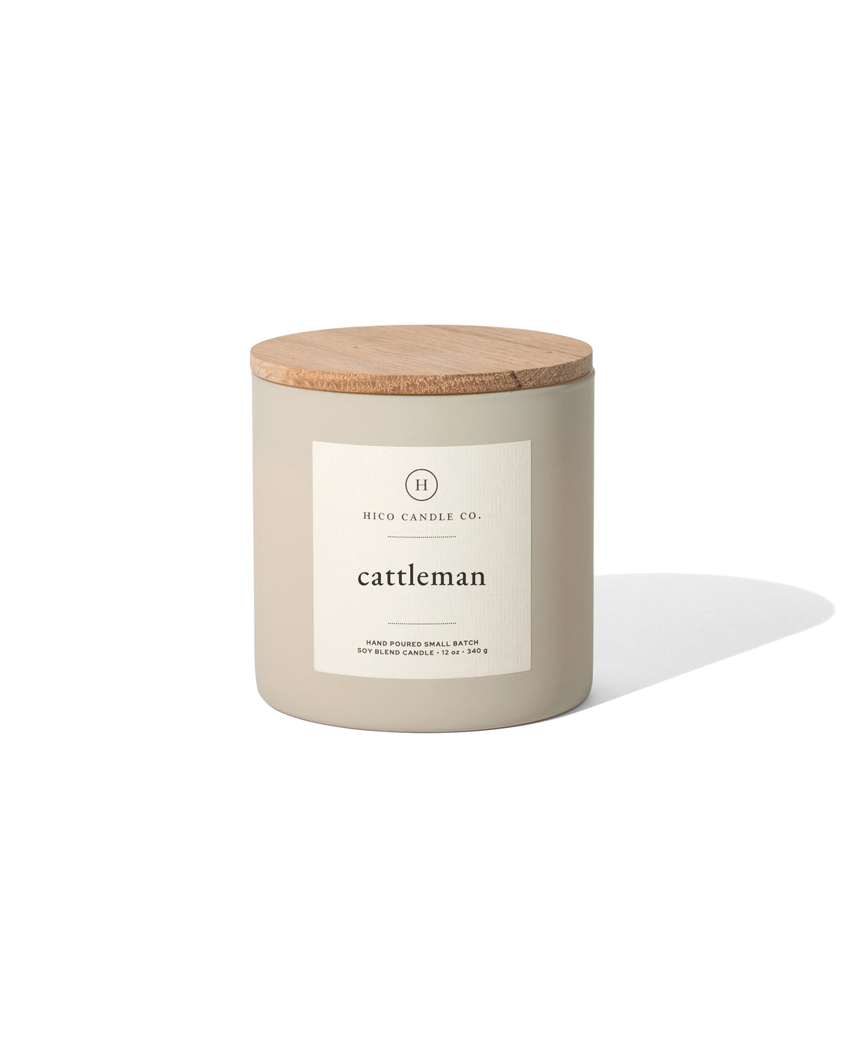 The 14oz. Cattleman candle by Faire features a beige jar with a wooden lid and minimalist label, hinting at its masculine soy fragrance. The jar casts a subtle shadow on a white background.
