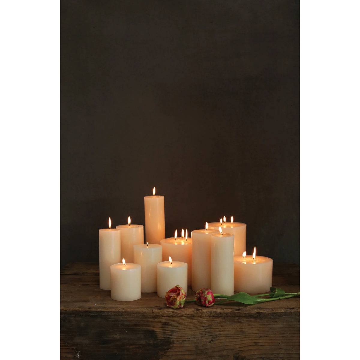 Creative Co-op Unscented Candle 4x4 candles in different heights are lit and placed on a rustic wooden surface, with two wilted red and yellow roses nearby against a dark background.