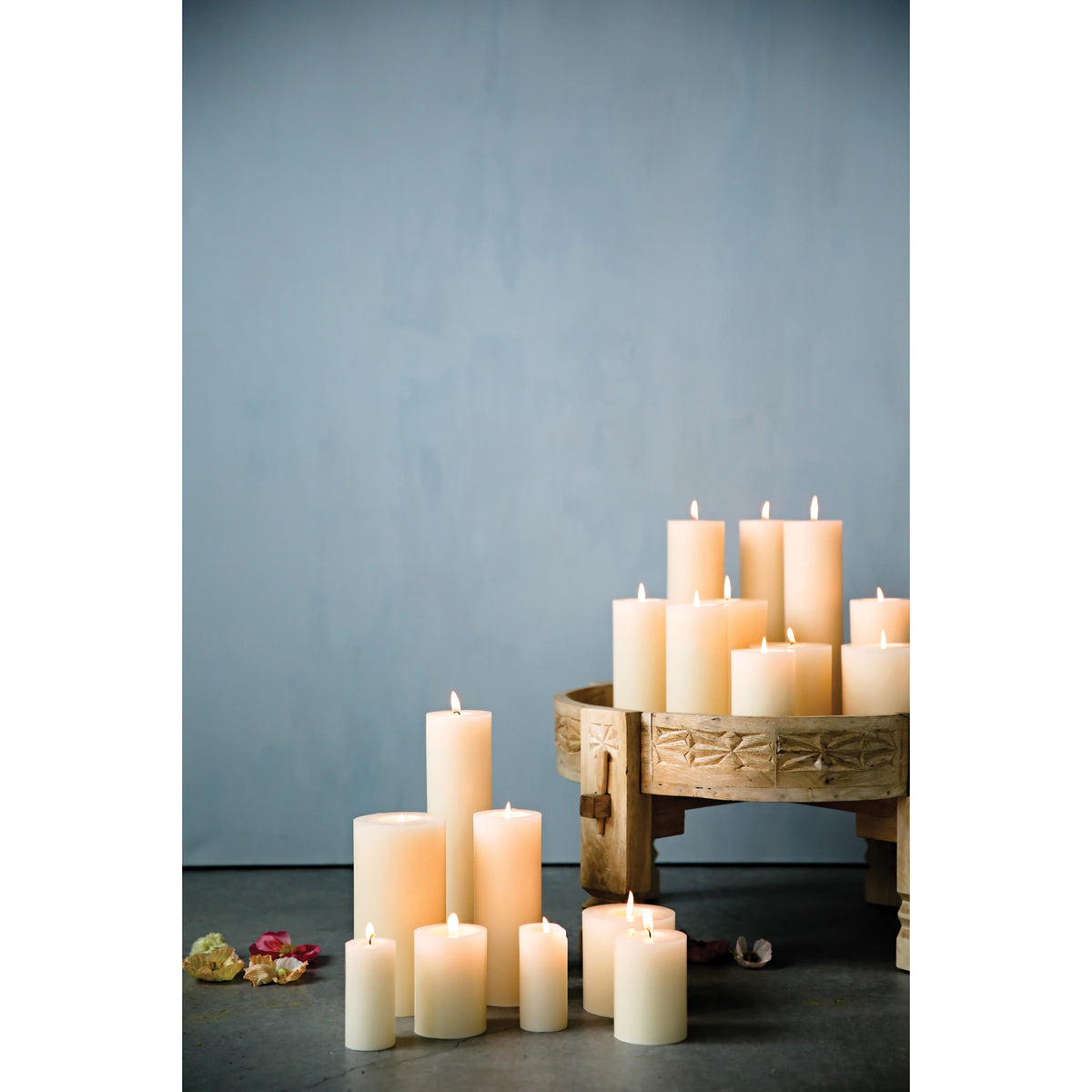 Creative Co-op Unscented Pillar Candles in varying heights, all lit, are arranged on the floor and a wooden pedestal with dried flowers nearby against a pale blue backdrop. Burn time: up to 145 hours.