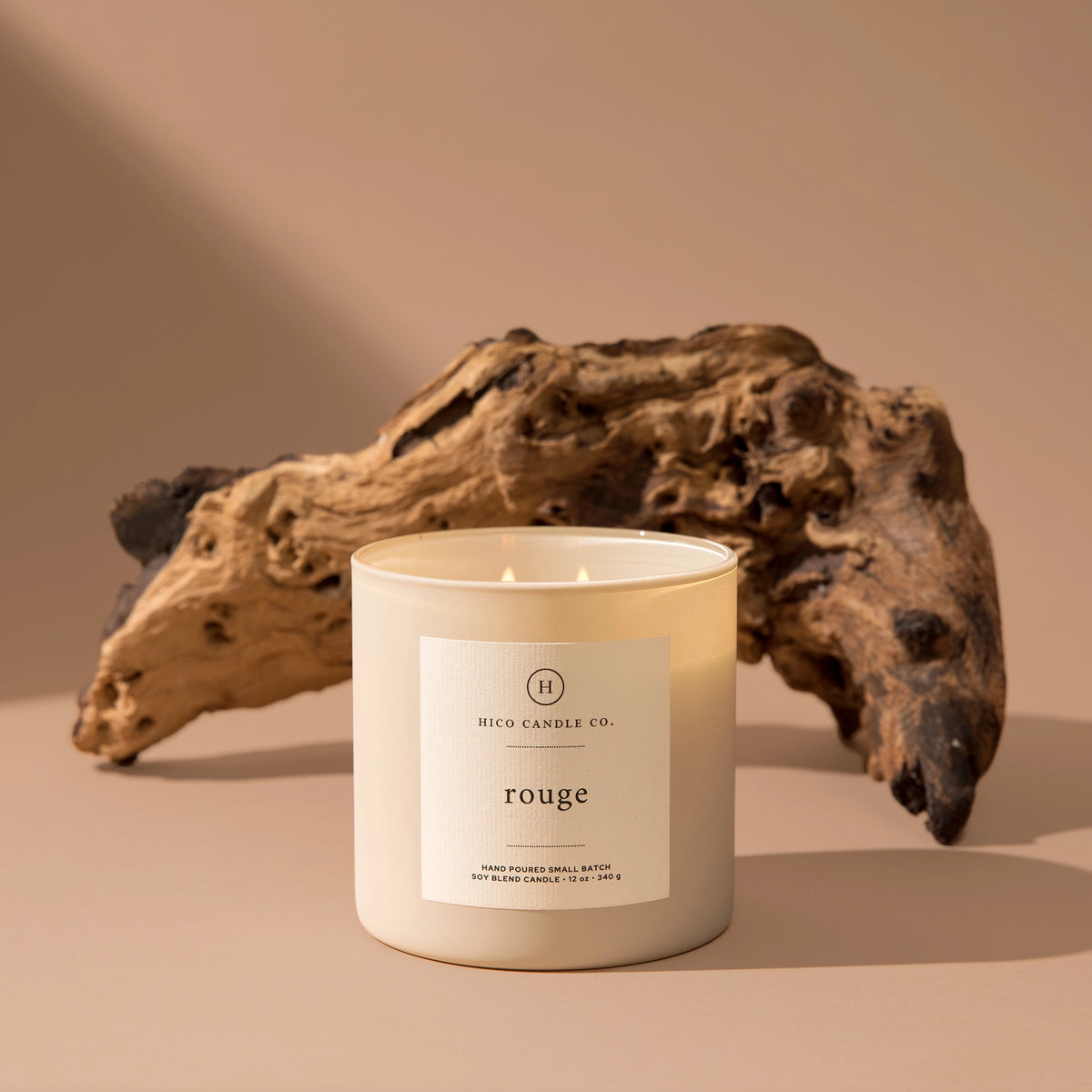 A hand poured 14oz. white luxury candle labeled "Rouge" from Faire rests on a beige surface with textured driftwood, evoking the sophisticated aroma of Baccarat Rouge 540 in a warm, neutral setting.