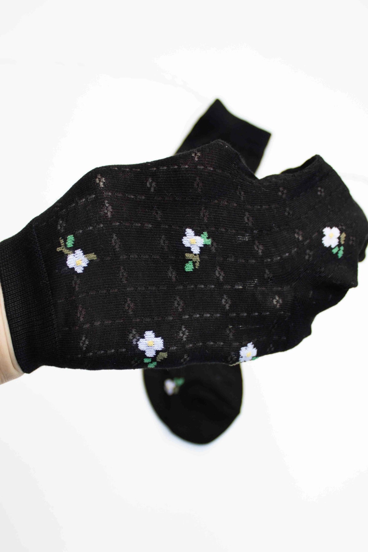 A hand displays a Faire Mini Floral Cotton Sock in black, featuring small white and yellow flowers; a matching sock is softly blurred in the background on a white surface.