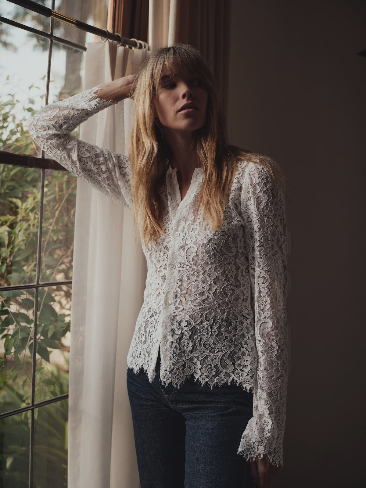 VERONA white floral lace button-down shirt paired with blue jeans, worn by a woman leaning against a window.