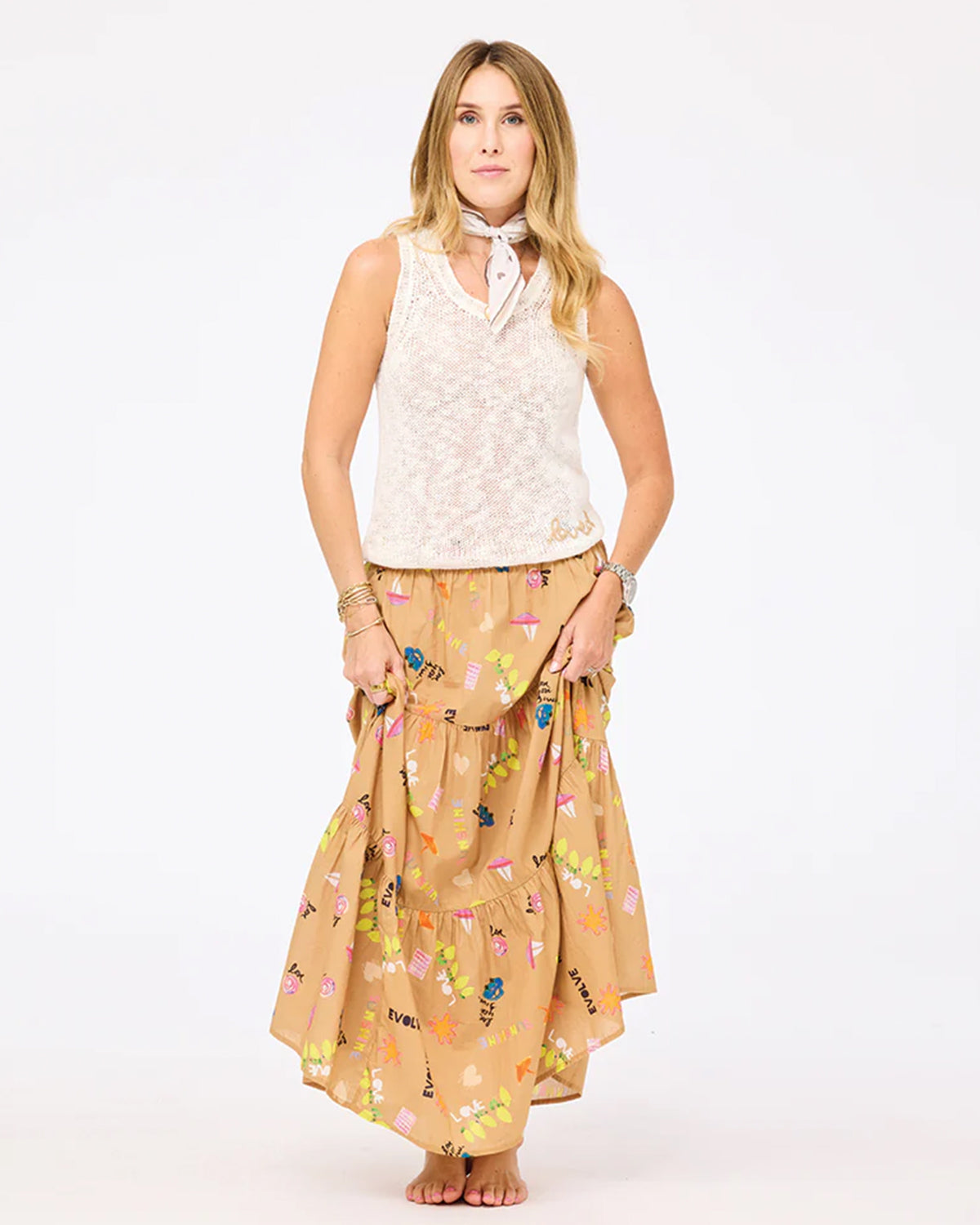 A woman stands barefoot against a white background, wearing the Kerri Rosenthal James Sweater Tank, a light tan floral maxi skirt with ruffles, several bracelets, and a white bandana tied around her neck.