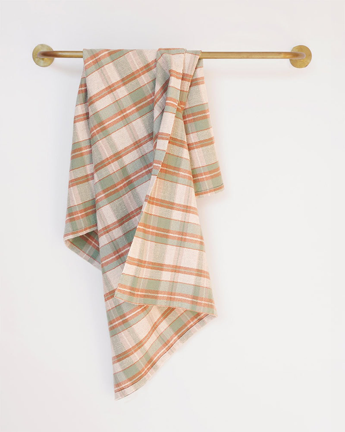 The Faire Cook's Cloth, a soft cotton-linen blend kitchen towel in muted green, orange, and beige plaid, hangs neatly over a brass towel bar on a plain white wall, combining style with durability.