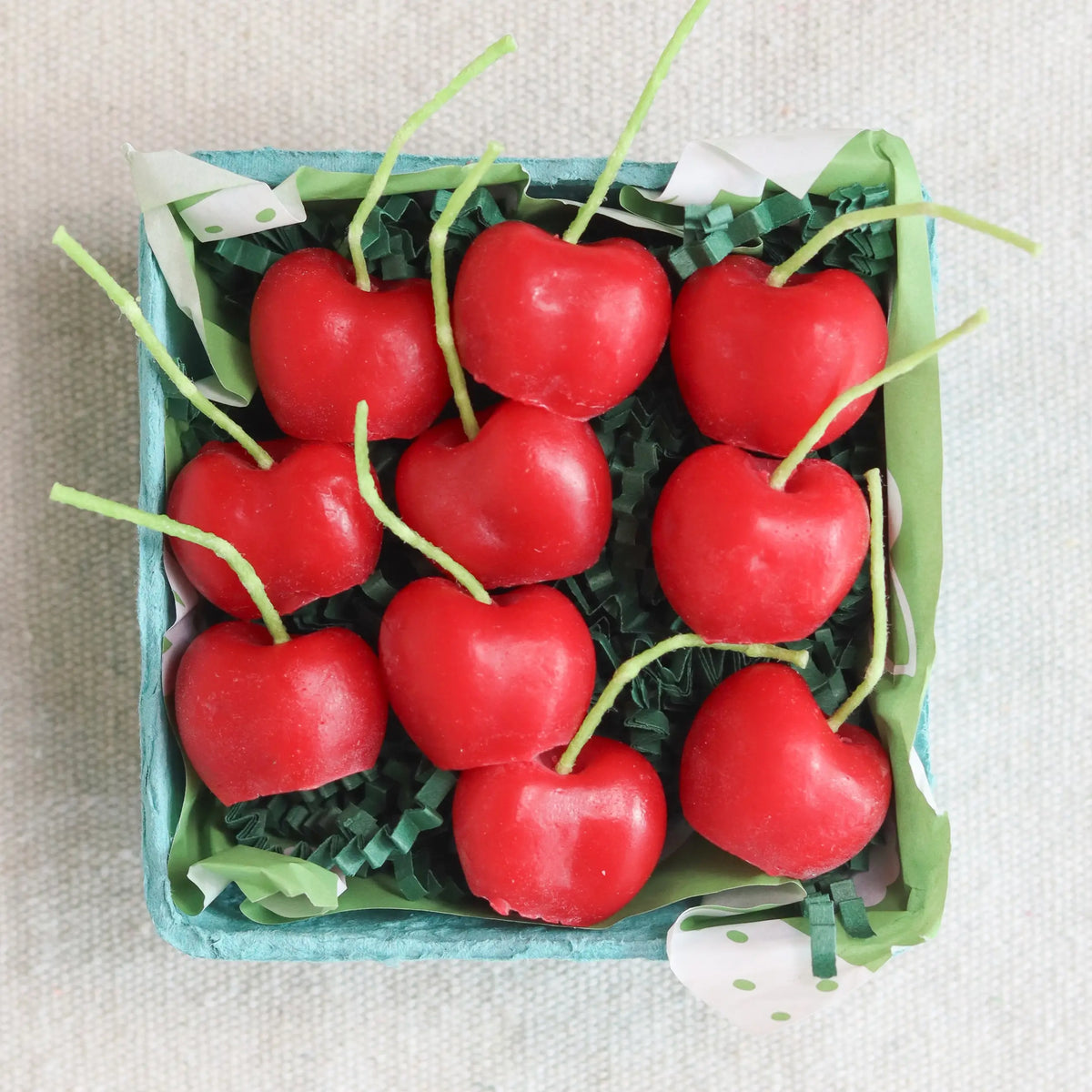 The Cherry Beeswax Candles by Faire come in a small blue container with nine heart-shaped, red cherry-like candles nestled on green paper shreds.