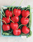 The Cherry Beeswax Candles by Faire come in a small blue container with nine heart-shaped, red cherry-like candles nestled on green paper shreds.