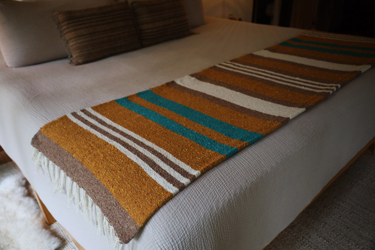 Handwoven striped blanket in brown, gold, teal, and white on bed; Bungalow Furniture & Accessories.