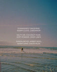 A person stands in shallow ocean waves under a blue sky. Overlaid text lists the fragrance notes of Faire’s SUNBURN 12 oz candle: sunwashed tangerine, warm clove, crisp linen, salty air, sandalwood, and sheer musk.