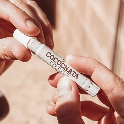 Two hands hold a tube of Faire Cocochata Lip Balm, handcrafted with organic coconut oil, as sunlight brightens the scene against a softly blurred neutral background.