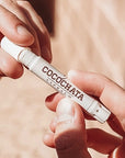 Two hands hold a tube of Faire Cocochata Lip Balm, handcrafted with organic coconut oil, as sunlight brightens the scene against a softly blurred neutral background.