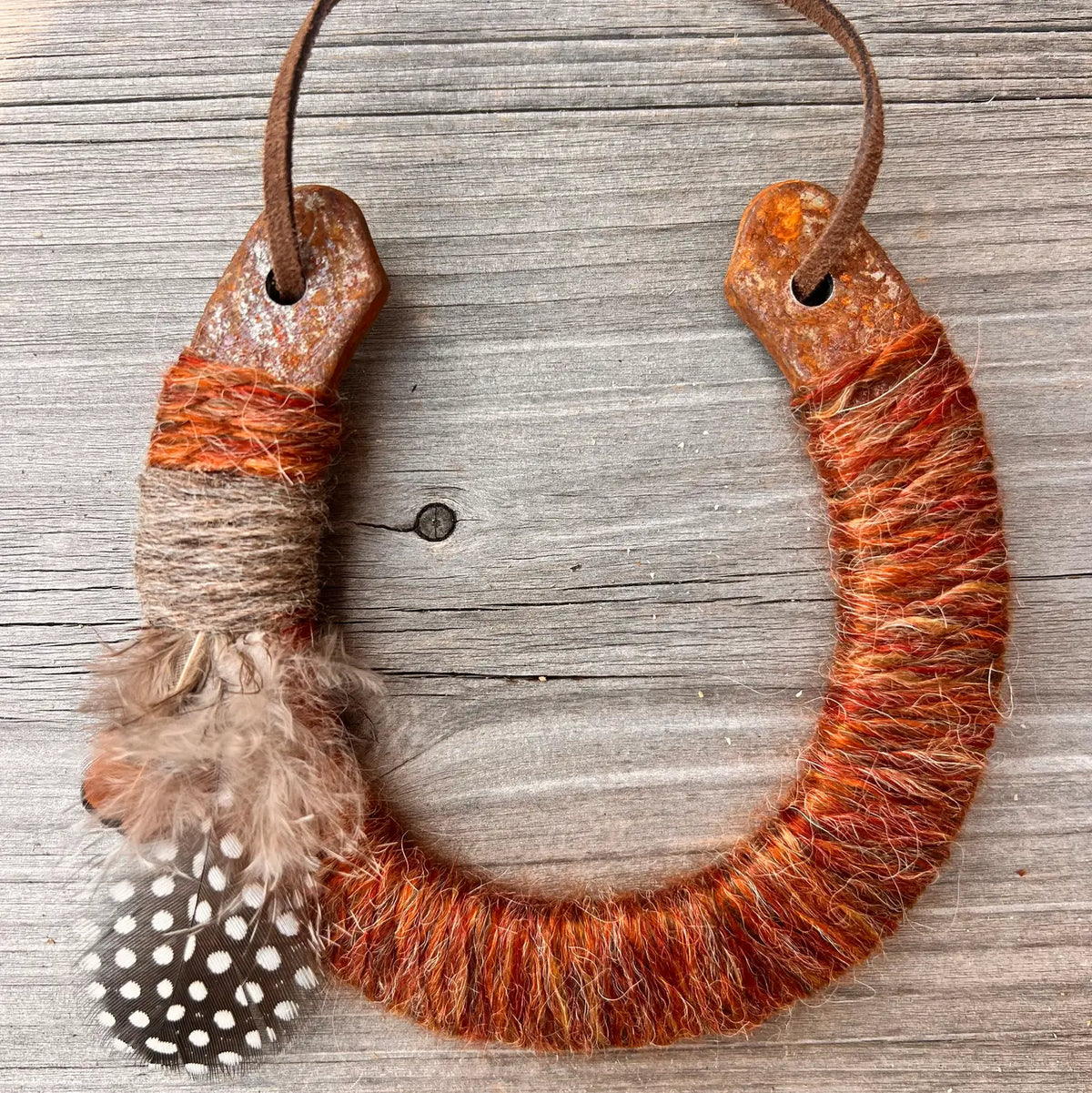 The Lucky Horseshoe by Faire is an upcycled keepsake wrapped in orange-brown yarn and twine, accented with a fluffy brown feather and a spotted feather, displayed on weathered wood.
