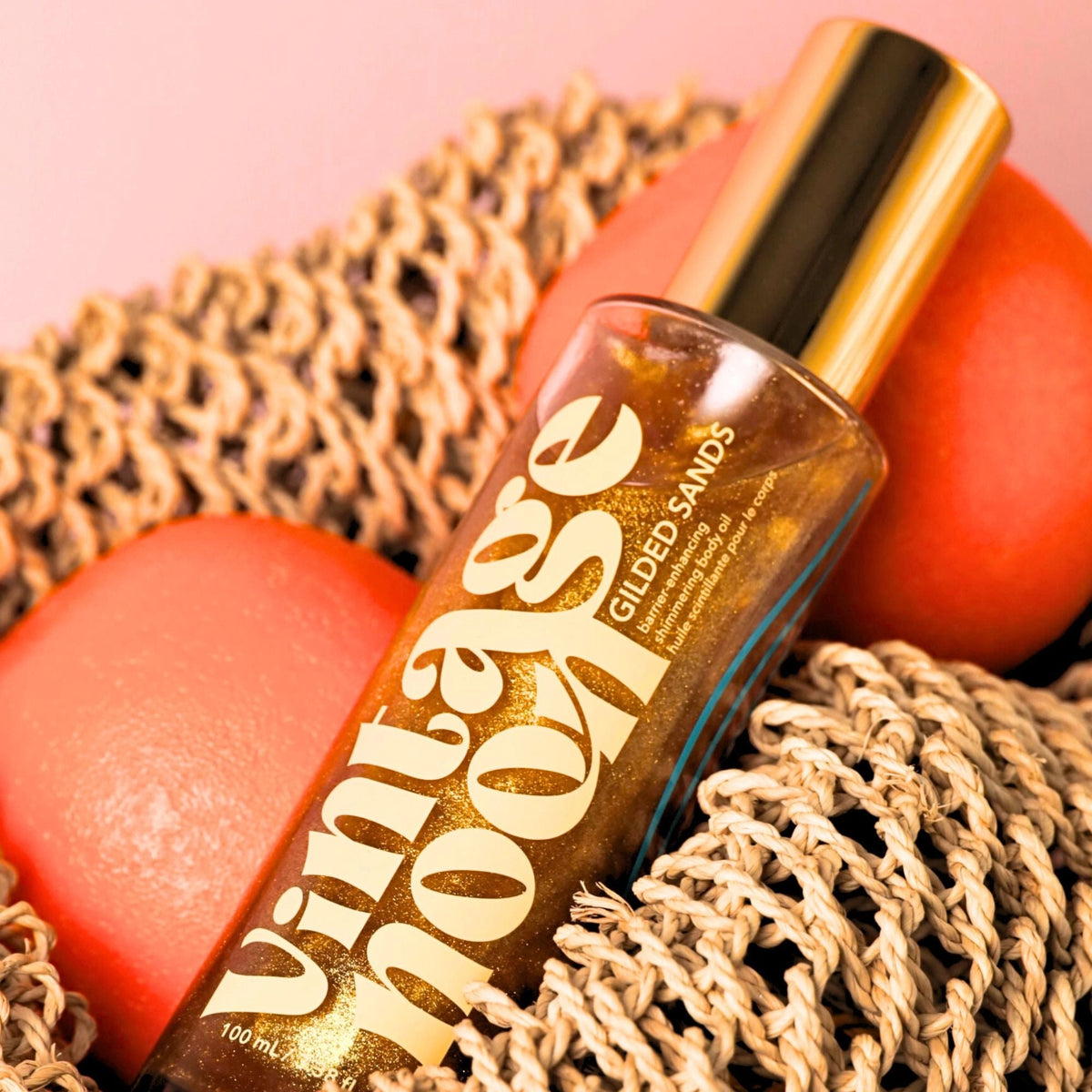 A bottle of Faire Shimmer Body Oil with gold lettering sits on a woven mat, flanked by two coral-colored stones against a soft pink background.