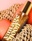 A bottle of Faire Shimmer Body Oil with gold lettering sits on a woven mat, flanked by two coral-colored stones against a soft pink background.