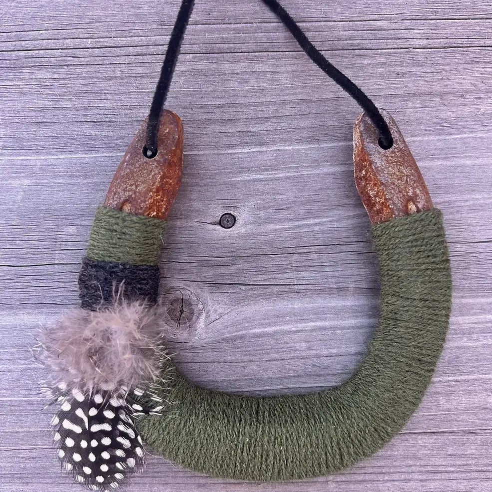 The Faire Lucky Horseshoe features an upcycled horseshoe wrapped in olive green yarn with grey and white spotted feathers on one side, hanging from a black cord on weathered wood. A unique housewarming gift for any space.