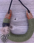The Faire Lucky Horseshoe features an upcycled horseshoe wrapped in olive green yarn with grey and white spotted feathers on one side, hanging from a black cord on weathered wood. A unique housewarming gift for any space.