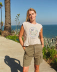 A woman wearing the Faire Hotel Luna Muscle Tank and olive green shorts stands outdoors by the ocean, surrounded by palm trees, lush plants, and a clear blue sky.