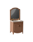 The Maileg Sink Dresser is a small brown dollhouse bathroom sink featuring a gold faucet, white countertop, cabinet door, and an attached rectangular mirror with rounded edges, shown against a plain white background.