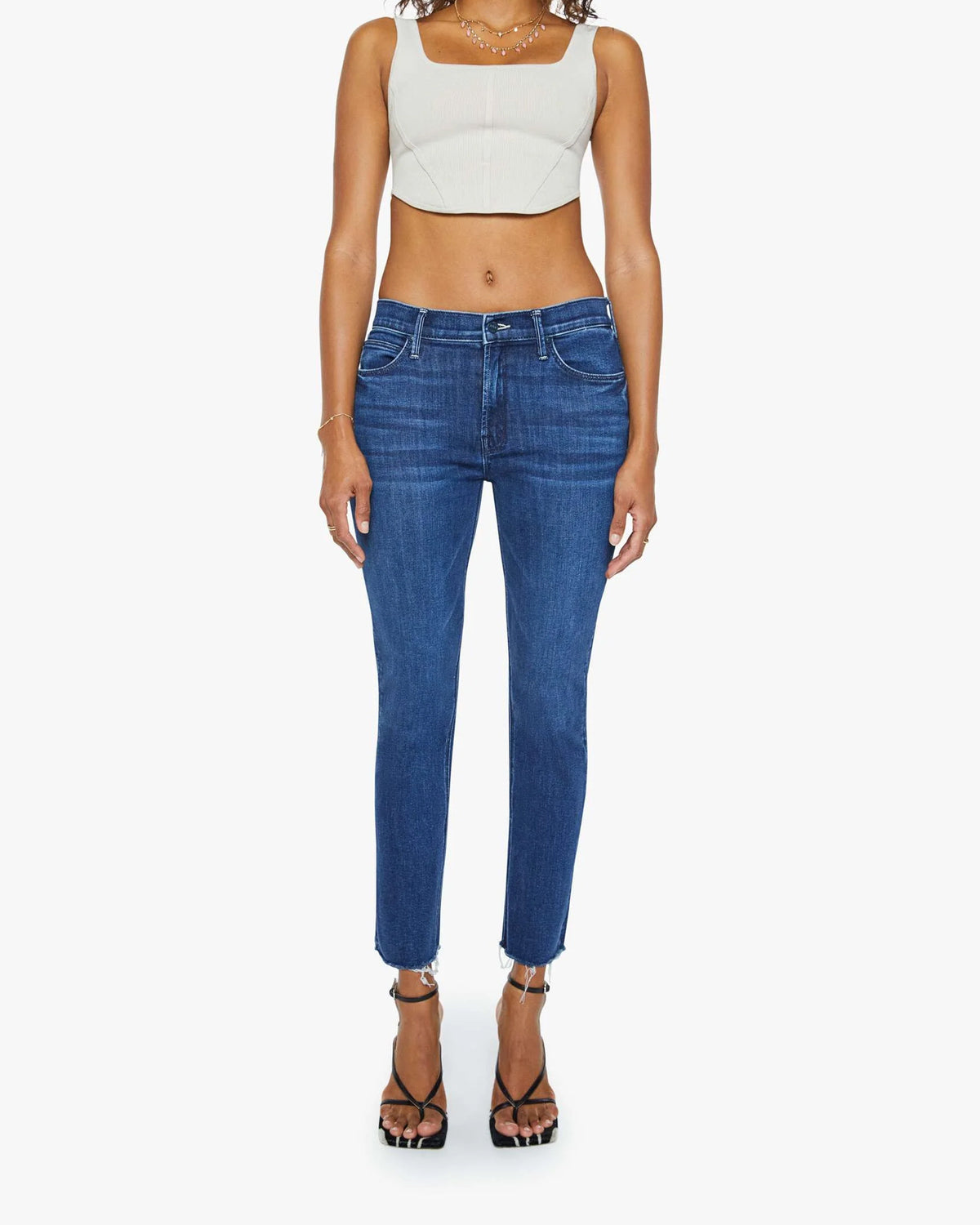 A person stands facing forward in a beige crop top and Mother’s Mid Rise Dazzler Ankle Fray blue jeans, paired with black heeled sandals, against a plain white background.